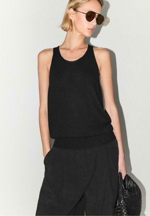 Kvinde med kort blondt hår iført mørke solbriller, sort ærmeløs top, sorte bukser og holder en tekstureret sort taske mod en ensfarvet baggrund.