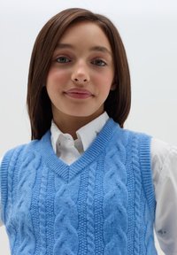 Hellblauer gestrickter Pullunder mit Zopfmuster, mit V-Ausschnitt und geripptem Kragen, getragen über einem weißen Hemd mit Kragen.