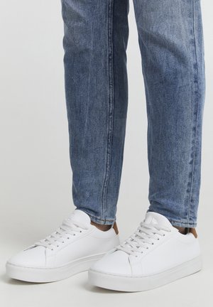 Weiße Leder-Sneaker mit flachen Sohlen, runden Zehen und Schnürdesign. Hellbrauner Akzent an der Ferse. Getragen mit blauen Jeanshosen.