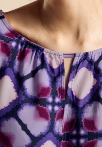 Blusa viola con scollatura rotonda, bordino arricciato e dettaglio a occhiale. Il tessuto ha una consistenza morbida con motivi astratti multicolori.