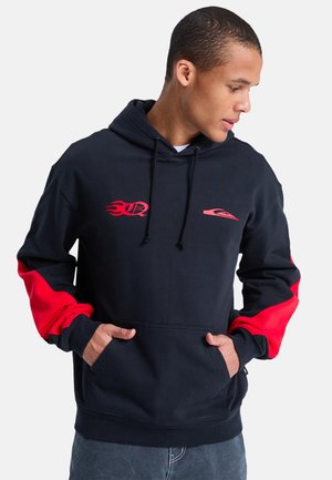 Młody mężczyzna w czarnej bluzie z kapturem z czerwonymi logo płomieni i fal, czerwonymi łatami na rękawach, z rękami w przedniej kieszeni, patrzący w prawo.