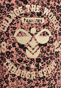 Rosa tyg med svart blommönster, med guldglittrande text som säger "FÖRÄNDRA VÄRLDEN GENOM SPORT" och en logotypdesign.