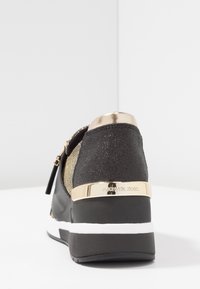 Zwarte en gouden sneaker met een gestructureerde bovenkant, metallic accenten, zichtbare ritssluiting en een wigzool met een witte streep en branding.