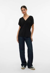 Donna giovane con capelli scuri, indossando un top pieghettato nero a V, jeans blu scuro e sneakers, in piedi su uno sfondo bianco.