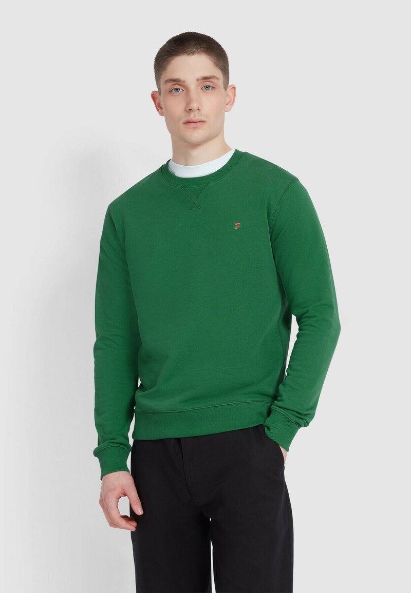 Groene katoenen sweater met een ronde hals en ribgebreide manchetten, met een klein logo op de borst, gedragen met zwarte pants.