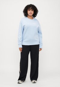 Helles blaues Sweatshirt mit gerippten Bündchen und Saum, mit weißem gesticktem Text. Kombiniert mit schwarzen weiten Hosen und Turnschuhen.