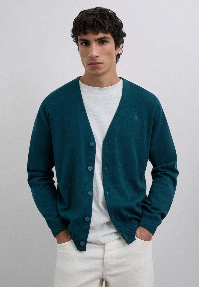 Cardigan teal din material moale, cu guler în V, șase butoane, manșete cotate și o croială lejeră, purtat peste un tricou gri deschis.