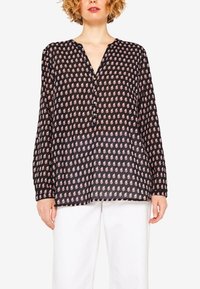 Frau trägt eine schwarze Langarmbluse mit rosa Paisley-Muster und weiße Hosen, steht vor einem weißen Hintergrund.