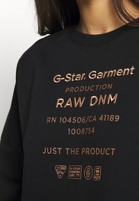 Felpa nera con testo ricamato color rame che include il nome del marchio, informazioni di produzione e loghi. Tessuto liscio, scollo rotondo.