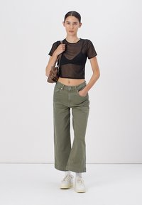 Top corto nero trasparente, pantaloni larghi verde oliva e sneakers bianche. L'outfit include una piccola borsa marrone. Design semplice e moderno.