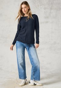 Lange Ärmeloberteil aus Navy-Wollmischung mit gerippter Textur, kombiniert mit hellblauen weit geschnittenen Jeans und weißen Sneakers. Einfaches Design, lässige Passform.