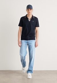 Camisa polo negra con cuello, jeans de mezclilla azul claro y zapatillas blancas. La camisa tiene un detalle a rayas cerca del cuello. Se lleva una gorra gris.