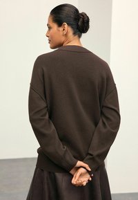 Nainen, jolla on hiukset nutturalla, pukeutuneena tummanruskeaan pitkähihaiseen neuleeseen ja hameeseen, seisoo kädet ristissä selän takana.