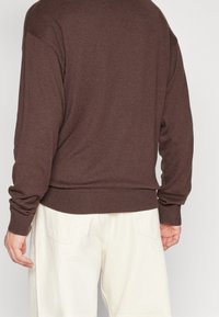 Mężczyzna w brązowym swetrze z długim rękawem i ecru spodniach, widziany z tyłu w zrelaksowanej pozycji.