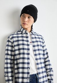 Mann trägt eine schwarze Strickmütze und ein kariertes Hemd in Blau-Weiß über einem weißen T-Shirt mit Logo und schaut nach links vor einem schlichten weißen Hintergrund.
