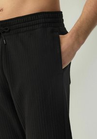 Pantalones de tela negra con relieve, cintura elástica y bolsillo lateral. La textura es suave con líneas verticales.