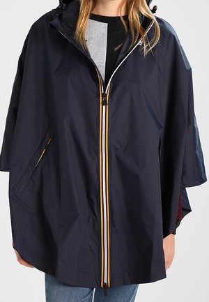 Personne portant un poncho de pluie bleu marine avec une fermeture éclair rayée jaune et blanche et une capuche, par-dessus une chemise noire et grise.