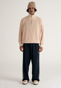 Man in een beige gebreide muts en een ritsvest met donkere wijde broek en beige schoenen, staande tegen een effen achtergrond.
