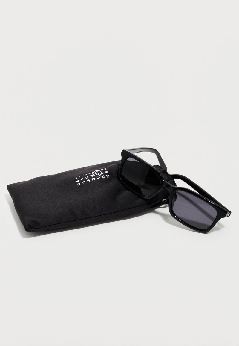 Occhiali da sole rettangolari neri poggiati su una custodia morbida nera con logo numerico bianco, su una superficie bianca.