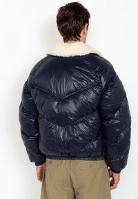 Marineblauwe gewatteerde jas met een schapenvachtkraag. Voorzien van een doorgestikt patroon en een elastische zoom. Gladde, glanzende textuur; ritsluiting niet zichtbaar.