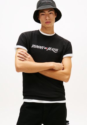 Νέος άνδρας που φοράει μαύρο t-shirt Tommy Jeans και καπέλο κάπα, με σταυρωμένα χέρια, μπροστά σε απλό λευκό φόντο.