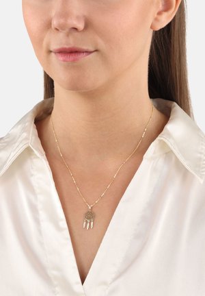 Collana dorata con un pendente a forma di cattura sogni, caratterizzato da un design rotondo e tre piume pendenti, indossata su una camicia bianca con colletto.