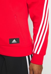 Sudadera roja con tres franjas blancas horizontales en las mangas, bolsillo con cremallera y un parche negro del logo de Adidas en el dobladillo.
