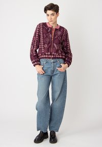 Veste bomber bordeaux avec broderies florales, poignets et ourlet côtelés, associée à un jean baggy bleu clair et des chaussures à plateforme noires.