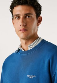 Felpa di cotone blu con scollo a costine, caratterizzata da un logo bianco "PEPE JEANS LONDON" sul petto, indossata sopra una camicia a righe con colletto.
