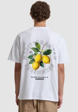 Personne portant un t-shirt blanc avec une illustration d'une branche de citronnier et le texte "Mediterranean Freshness" et "The Golden Lemon Groves of Sorrento" au dos.