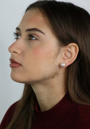 Jeune femme aux longs cheveux bruns et à la peau olive, portant des boucles d'oreilles en perle et un col roulé bordeaux, regardant vers la gauche.