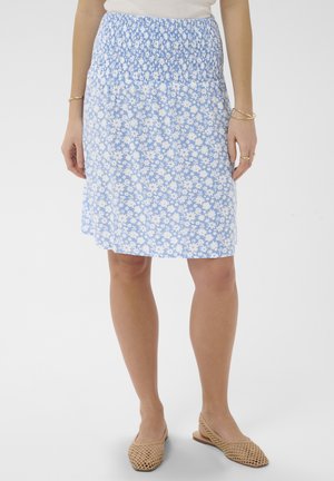 Kvinde iført lysblå blomster nederdel med smocket talje, hvid top, guld armbånd og beige vævede flade sko, på hvid baggrund.