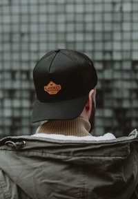 Fekete sapka hálós hátsó résszel, rajta egy barna bőrpánt a "BLACKSKIES" logóval. Bézs bordázott galléros dzsekivel viselve.