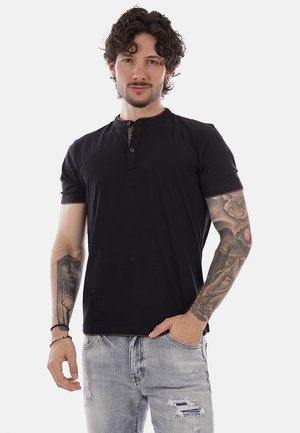 Uomo con capelli ricci che indossa una maglietta Henley nera a maniche corte e jeans chiari strappati, con le braccia tatuate e la mano in tasca.