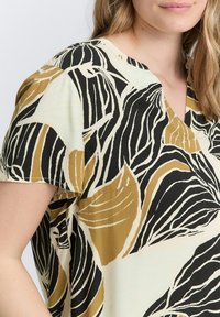 Lys stof bluse med V-hals, korte ærmer og et dristigt bladdesign i sort, crème og nuancer af guld.