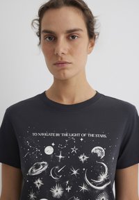 Dark gray cotton t-shirt featuring white celestial graphics, including stars, moons, and planets, with text "TO NAVIGATE BY THE LIGHT OF THE STARS."