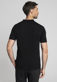 Mężczyzna z krótkimi brązowymi włosami, ubrany w czarną prążkowaną koszulę polo z krótkim rękawem, odwrócony z rękami w kieszeniach ciemnych spodni, na szarym tle.