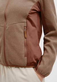 Brown jacket featuring a textured mesh fabric on the front with solid brown accents, a zippered pocket, and a smooth finish.