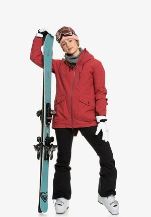 Veste de ski rouge, pantalon noir, gants blancs ; tenant des skis bleus avec fixations. Bonnet rose et lunettes de ski. Design moderne, tenue fonctionnelle.
