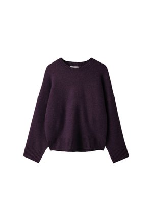 Jeune femme en pull violet foncé, jupe courte gris foncé, bottes noires montantes à lacets, tenant un sac à main gris, debout devant un fond gris.