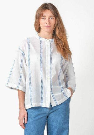 Junge Frau mit langen braunen Haaren, die ein lockeres weißes Hemd mit blauen vertikalen Streifen und blaue Jeans vor einem schlichten weißen Hintergrund trägt.