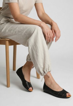 Persoon zit op een houten stoel, draagt beige broek en zwarte open schoenen met uitgesneden patronen, handen rusten op de knie.