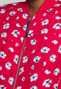 Veste bomber rouge avec une fermeture éclair, présentant un motif floral de fleurs blanches et bleues. Fabriquée en tissu lisse et léger avec des bordures côtelées.