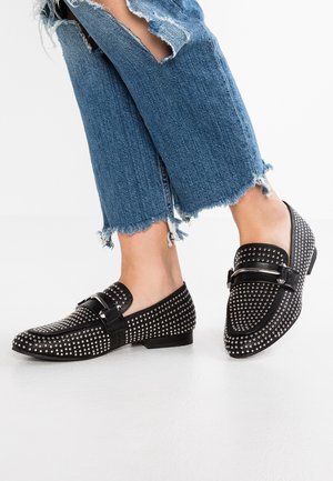 Fødder iført sorte loafers med sølvnitter kombineret med blå cropped jeans med frynset kant og revner på hvid baggrund.