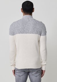 Jersei de punto con una textura de canalé, que presenta una sección superior gris y una sección inferior crema, cuello alto y pequeños detalles en los hombros.