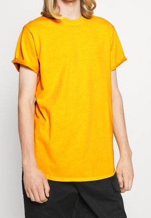 T-shirt en coton orange avec manches retroussées et col rond. Couleur unie avec une texture subtile. Porté avec un pantalon cargo noir.