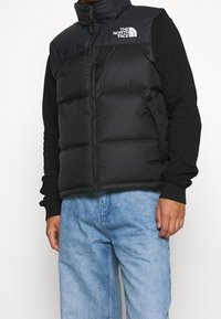 Gilet nero imbottito con collo alto, dotato di chiusura frontale con zip e logo ricamato sulla parte superiore sinistra. Indossato sopra una maglia nera a maniche lunghe.