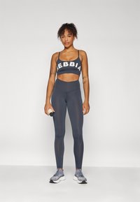 Dark gray sports bra with white "NEBBIA" logo, paired with high-waisted dark gray leggings. Texture appears smooth and stretchy; model holds a dumbbell.
