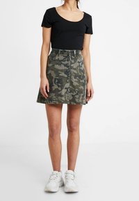 Kvinna som bär en svart T-shirt med rundad halsringning, en knappframskjortad camouflageskjol och vita sneakers, stående mot en enfärgad vit bakgrund.