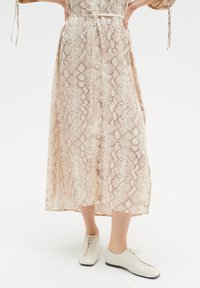 Nainen, joka on pukeutunut vaaleanbeigeeseen käärmeennahkakuosisessa midihameeseen, jossa on etupainikkeet, ja valkoisiin nauhakenkiin, seisoo kädet lanteilla.